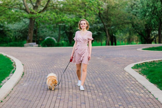 Atractiva mujer joven con perro spitz afuera y sonriendo a la cámara, caminando en el parque. Concepto de amistad entre personas y animales.