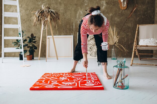 Foto atractiva mujer joven de pie en el piso con los pies descalzos y pintar con pincel y color blanco sobre lienzo rojo