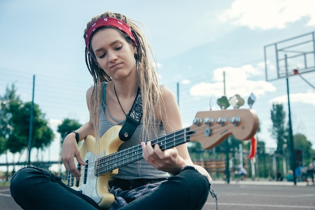 Atractiva mujer joven con rastas sentado en el campo de deportes y mirando concentrado mientras toca la guitarra