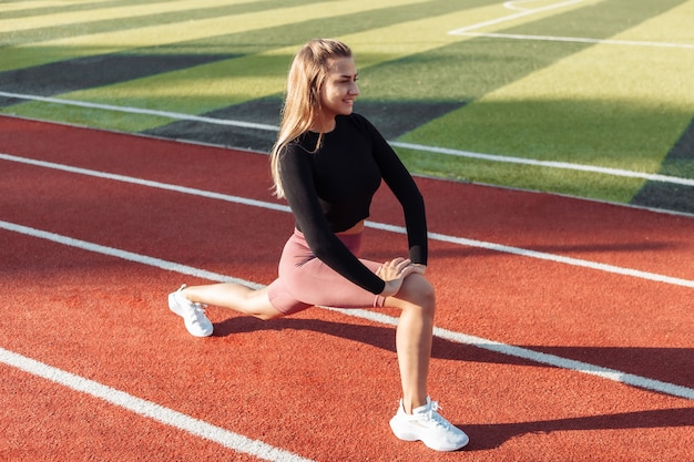 Atractiva mujer joven en ropa deportiva haciendo calentamiento previo al entrenamiento, estirando las piernas en el estadio. Concepto de estilo de vida saludable. Entrenamiento al aire libre