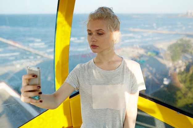 Atractiva mujer joven rubia haciendo selfie en su teléfono inteligente sentado en el carrusel