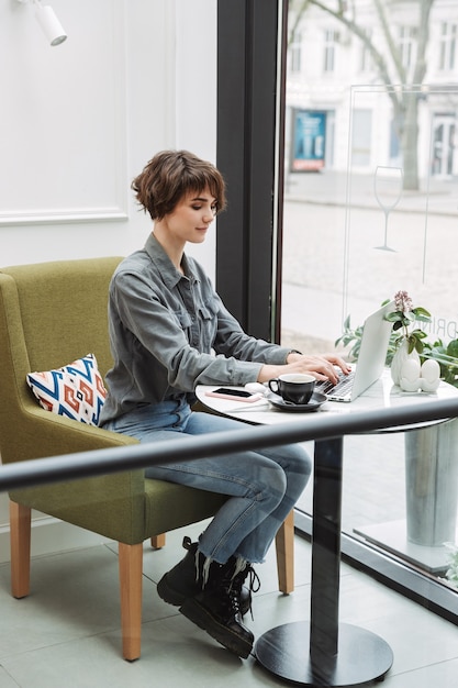 Atractiva mujer joven sentada en la mesa de café en el interior, trabajando en la computadora portátil, analizando documentos