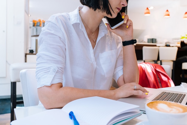 Atractiva mujer joven sentada en la oficina de coworking y escribiendo en su computadora portátil con teclado y tomando notas en la mañana. Escritorio con teléfono, cuaderno, vasos y una taza de café. Concepto de negocio