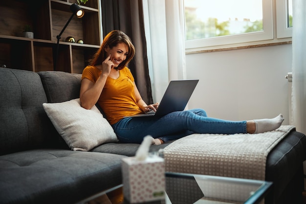 Atractiva mujer joven sentada en el sofá y usando su computadora portátil en casa.