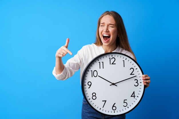 Atractiva mujer joven sosteniendo un gran reloj de pared contra el fondo azul.