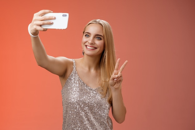 Atractiva mujer joven tierna rubia de 25 años en elegante vestido plateado tomando selfie extender brazo hacia arriba mostrar gesto de paz smartphone pantalla grabar video saludo a los fanáticos de internet, de pie fondo rojo