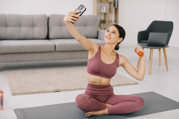 Atractiva mujer joven tomando selfie mostrando bíceps con mancuerna en casa