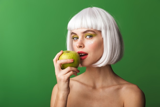 Atractiva mujer joven en topless con el pelo corto y blanco que se encuentran aisladas, comiendo una manzana