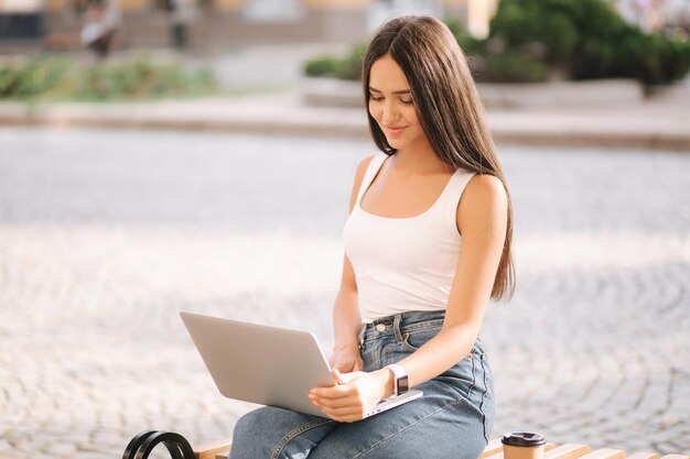 Atractiva mujer joven usa la computadora portátil afuera en el banco en verano Hermosa mujer morena