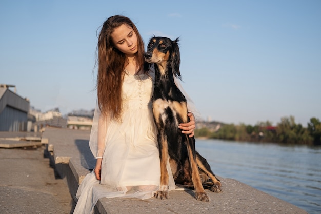 Atractiva mujer joven en vestido blanco sentada con perro saluki