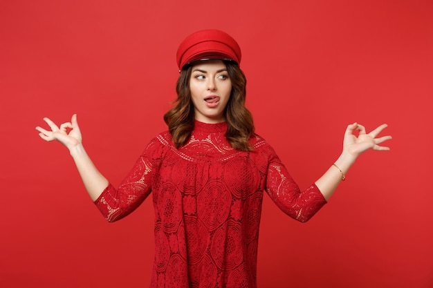 Atractiva mujer joven con vestido de encaje, gorra que muestra la lengua, mirando a un lado, tome las manos en un gesto de yoga relajándose aislado en un fondo rojo. Emociones sinceras de la gente, concepto de estilo de vida. Simulacros de espacio de copia.