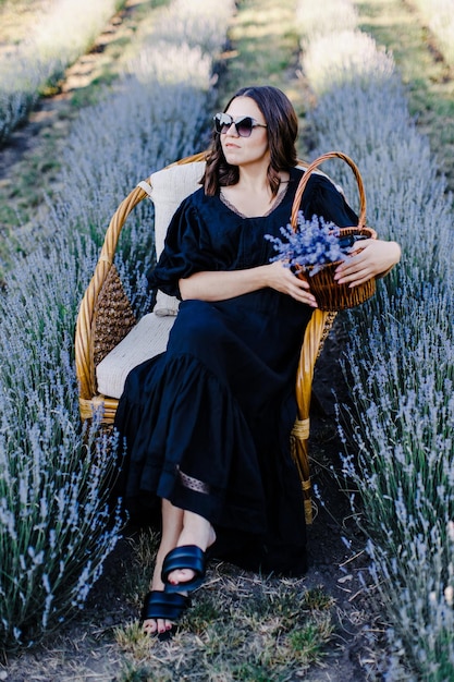 Atractiva mujer joven con vestido negro y gafas de sol sentada en una silla rodeada de un campo de lavanda y sosteniendo una canasta con flores