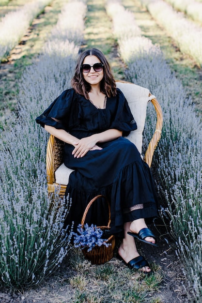 Atractiva mujer joven con vestido negro y gafas de sol sentada en una silla rodeada de un campo de lavanda