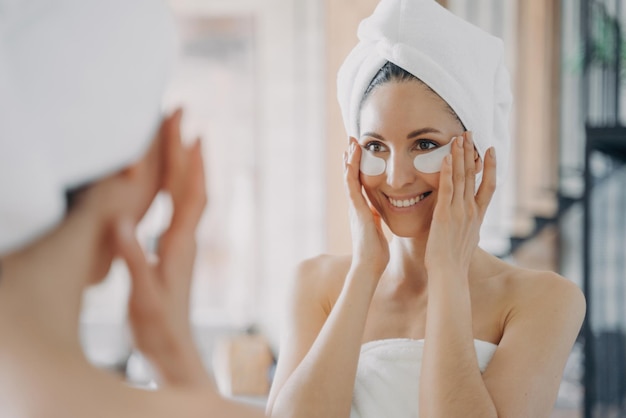 Foto atractiva mujer latina con parches debajo de los ojos mirando al espejo durante el tratamiento diario de cuidado de la piel