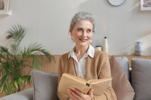 Atractiva mujer de mediana edad que disfruta leyendo un libro sentado en el sofá de su sala de estar sonriendo mientras lee.