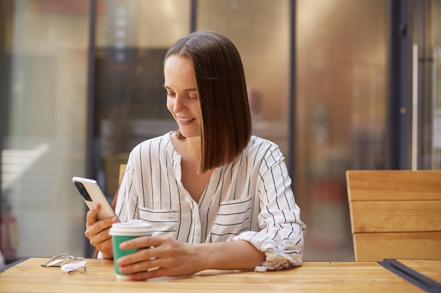 Atractiva mujer morena con atuendo formal mientras usa teléfonos inteligentes tomando café en un café disfrutando de un café sosteniendo un teléfono móvil leyendo noticias de Internet