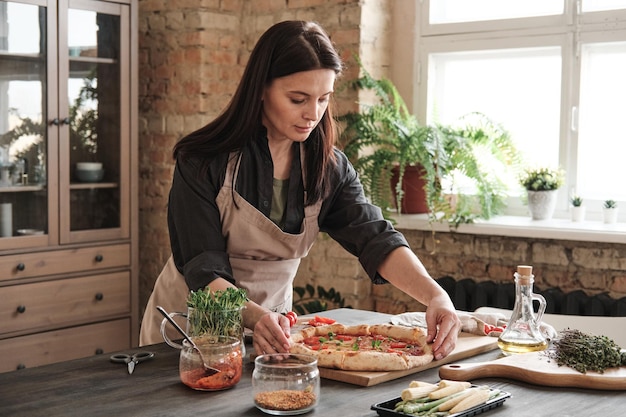 Atractiva mujer morena con delantal parada en el mostrador de la cocina y ajustando la pizza a bordo mientras la prepara para la cena