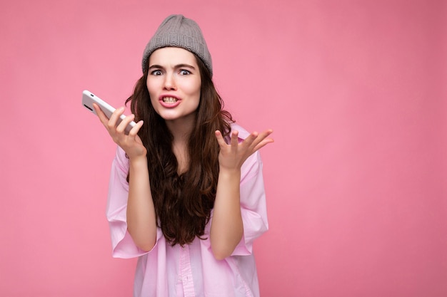 Atractiva mujer morena joven emocional con elegante camisa rosa y sombrero gris aislado sobre rosa