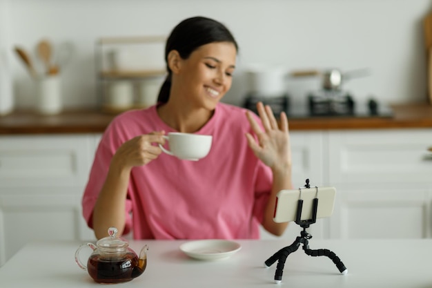 Atractiva mujer morena joven sonriente en camiseta rosa de gran tamaño está bebiendo té en la cocina Chica guapa feliz con sonrisa blanca como la nieve saluda a un amigo que se comunica por videollamada en línea en el teléfono móvil