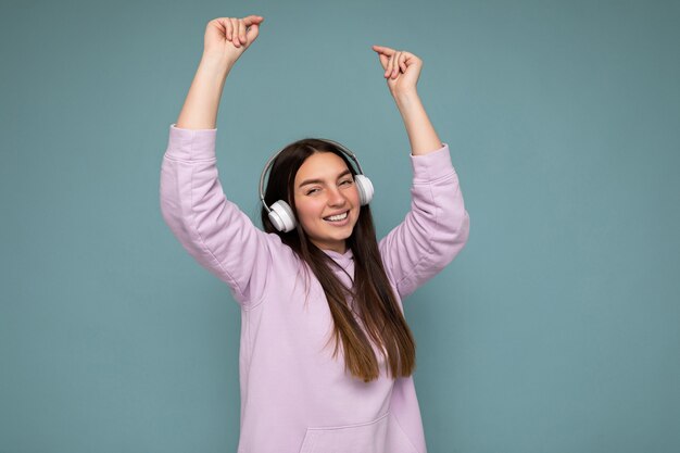 Atractiva mujer morena joven sonriente positiva con sudadera con capucha de color púrpura claro aislado sobre azul