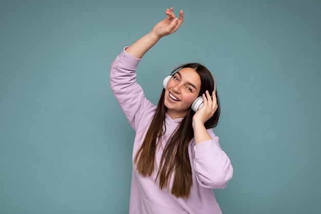 Atractiva mujer morena joven sonriente positiva vistiendo sudadera con capucha de color púrpura claro aislado sobre azul