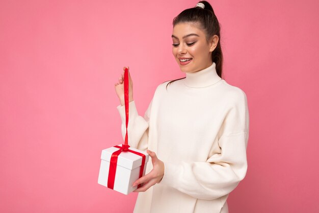 Atractiva mujer morena joven sorprendida positiva aislada sobre fondo rosa pared vistiendo un suéter blanco con caja de regalo y unboxing presente mirando la caja.