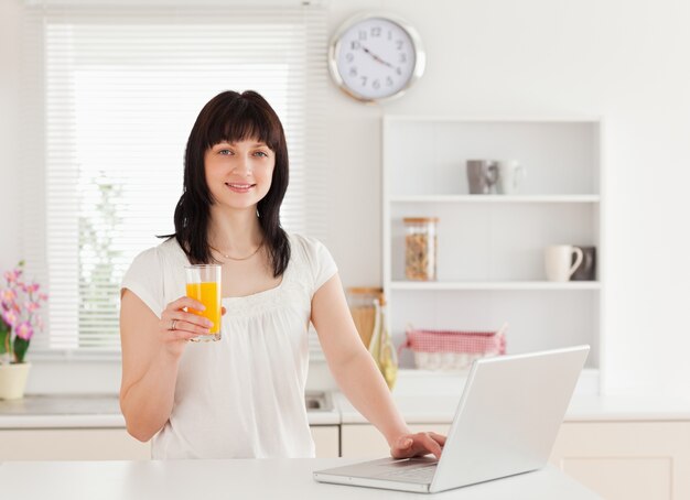 Atractiva mujer morena con un vaso de jugo de naranja mientras se relaja con su computadora portátil