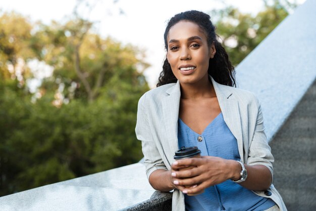 Atractiva mujer de negocios africana sonriente de pie al aire libre en la ciudad, apoyado en las escaleras, sosteniendo la taza de café para llevar