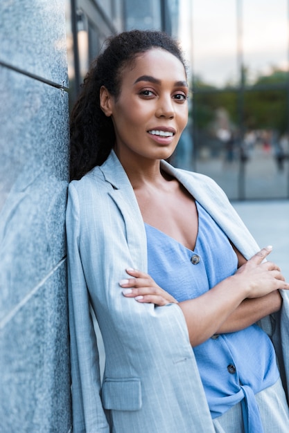 Atractiva mujer de negocios africana sonriente de pie al aire libre en la ciudad, apoyado en una pared