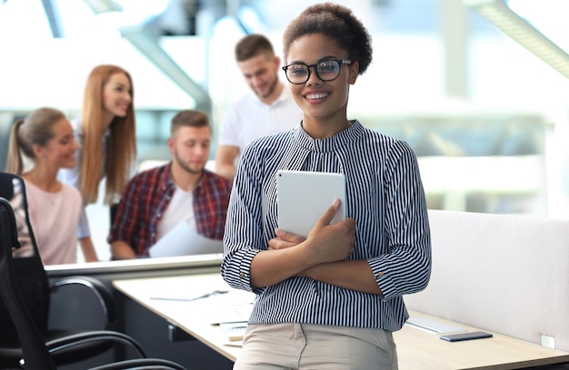 Atractiva mujer de negocios afroamericana mirando a la cámara y sonriendo mientras está de pie en la oficina