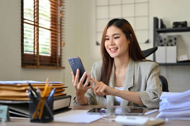 Atractiva mujer de negocios asiática en el escritorio de su oficina usando su teléfono inteligente para revisar su correo electrónico