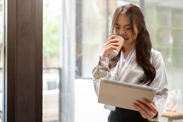 Una atractiva mujer de negocios asiática está tomando café y usando su tableta en un pasillo de oficina