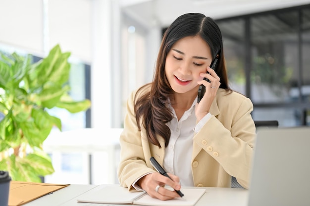 Atractiva mujer de negocios asiática tomando notas en su cuaderno mientras habla por teléfono