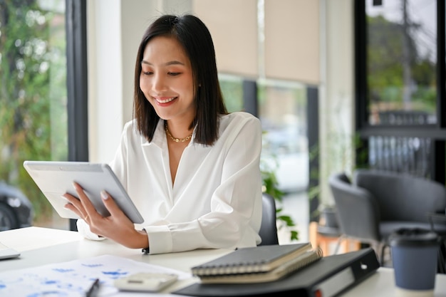 Atractiva mujer de negocios asiática usando su tableta para revisar su trabajo sentada en su escritorio en la oficina