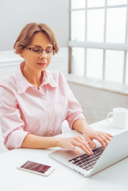 Atractiva mujer de negocios con camisa clásica y anteojos está usando una computadora portátil mientras trabaja en la oficina