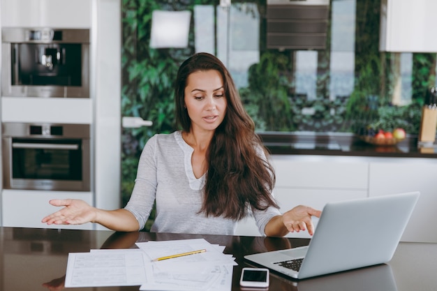 La atractiva mujer de negocios confundida que trabaja con documentos y una computadora portátil en la cocina de casa