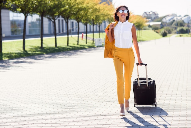 Atractiva mujer de negocios en elegante traje amarillo saca una maleta, se apresura al aeropuerto. Mujer de negocios atractiva va en un viaje de negocios tirando de su maleta detrás de ella.