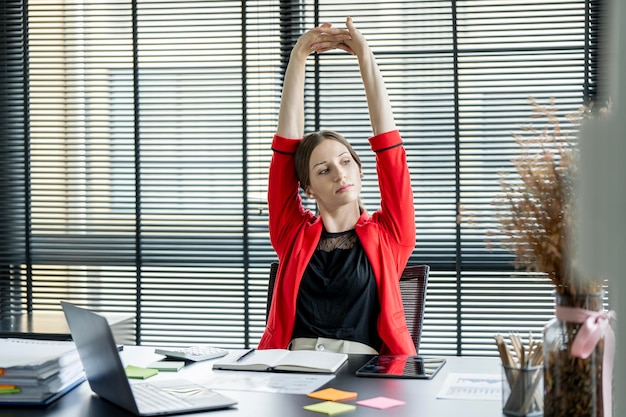 Atractiva mujer de negocios estirando las manos y el cuerpo tomando un descanso del trabajo en una laptop sentada en el escritorio y sonriendo mirando por la ventana