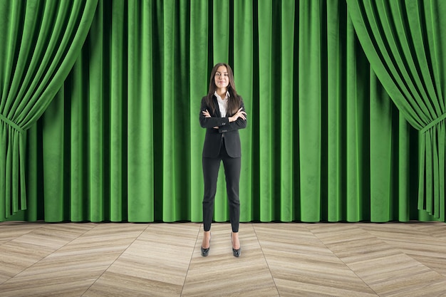 Atractiva mujer de negocios feliz con los brazos cruzados frente a la cortina verde y el suelo de madera Escenario de cine y teatro