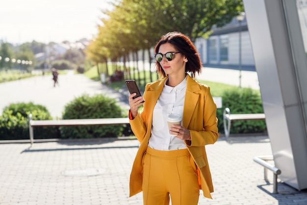 Atractiva mujer de negocios en gafas de sol y traje amarillo caminando cerca del centro de la oficina con una taza de café y utiliza smartphone. Concepto de estilo de vida y éxito empresarial. Almuerzo entre días laborables.