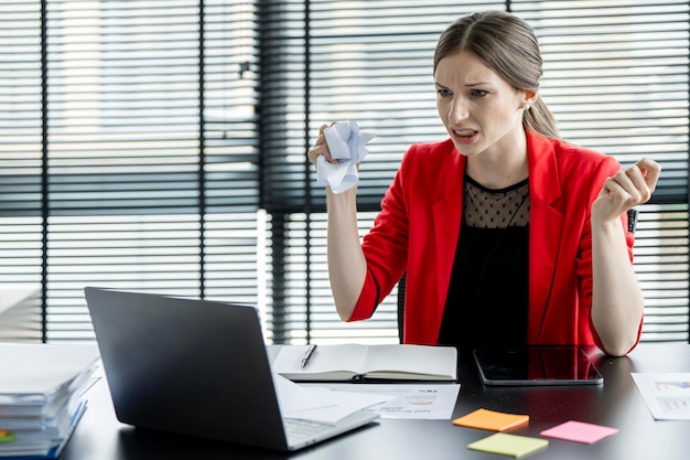 Atractiva mujer de negocios loca sentada en la oficina sintiéndose enojada, arrugando el papel y mirando la pantalla del portátil