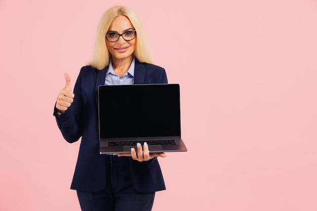 Atractiva mujer de negocios de mediana edad con anteojos sosteniendo una computadora portátil y mostrando la pantalla simulada en un fondo rosa