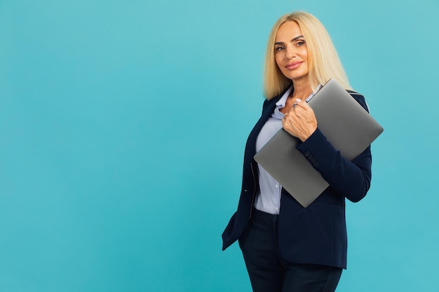 Atractiva mujer de negocios de mediana edad con gafas sosteniendo una laptop en un fondo azul