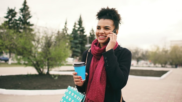 Atractiva mujer de negocios de raza mixta hablando de teléfonos inteligentes y bebiendo café camina en la calle de la ciudad