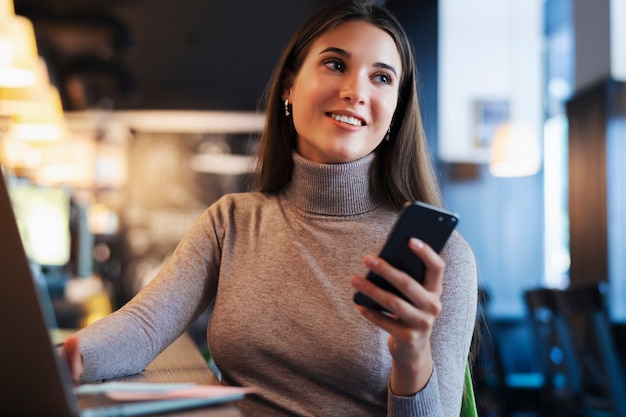 Atractiva mujer de negocios se sienta a la mesa frente a la computadora portátil y habla por teléfono móvil, negocia por teléfono.