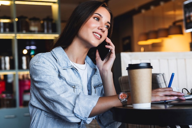 Foto atractiva mujer de negocios se sienta a la mesa frente a la computadora portátil y habla por teléfono móvil, negocia por teléfono.