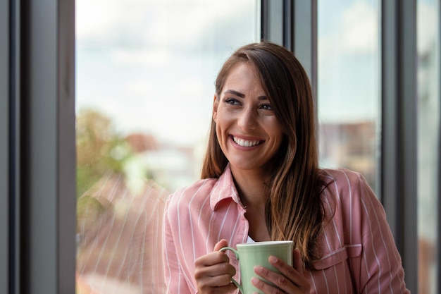 Atractiva mujer de negocios tomando un café mientras está parada en una ventana en una oficina con vista a la ciudad