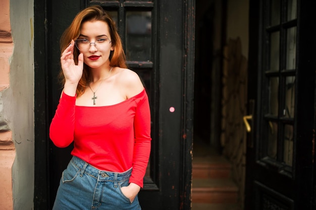 Atractiva mujer pelirroja con anteojos en blusa roja y falda de jeans posando en la calle contra la vieja puerta de madera