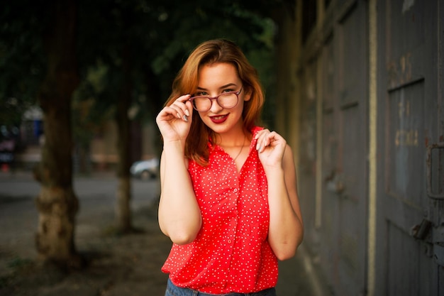 Atractiva mujer pelirroja con anteojos en blusa roja y falda de jeans posando