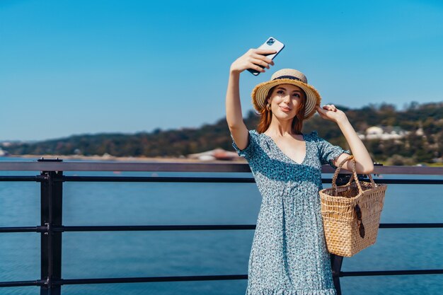 Atractiva mujer pelirroja posando en un día soleado haciendo selfie con teléfono camina por el muelle cerca del ...